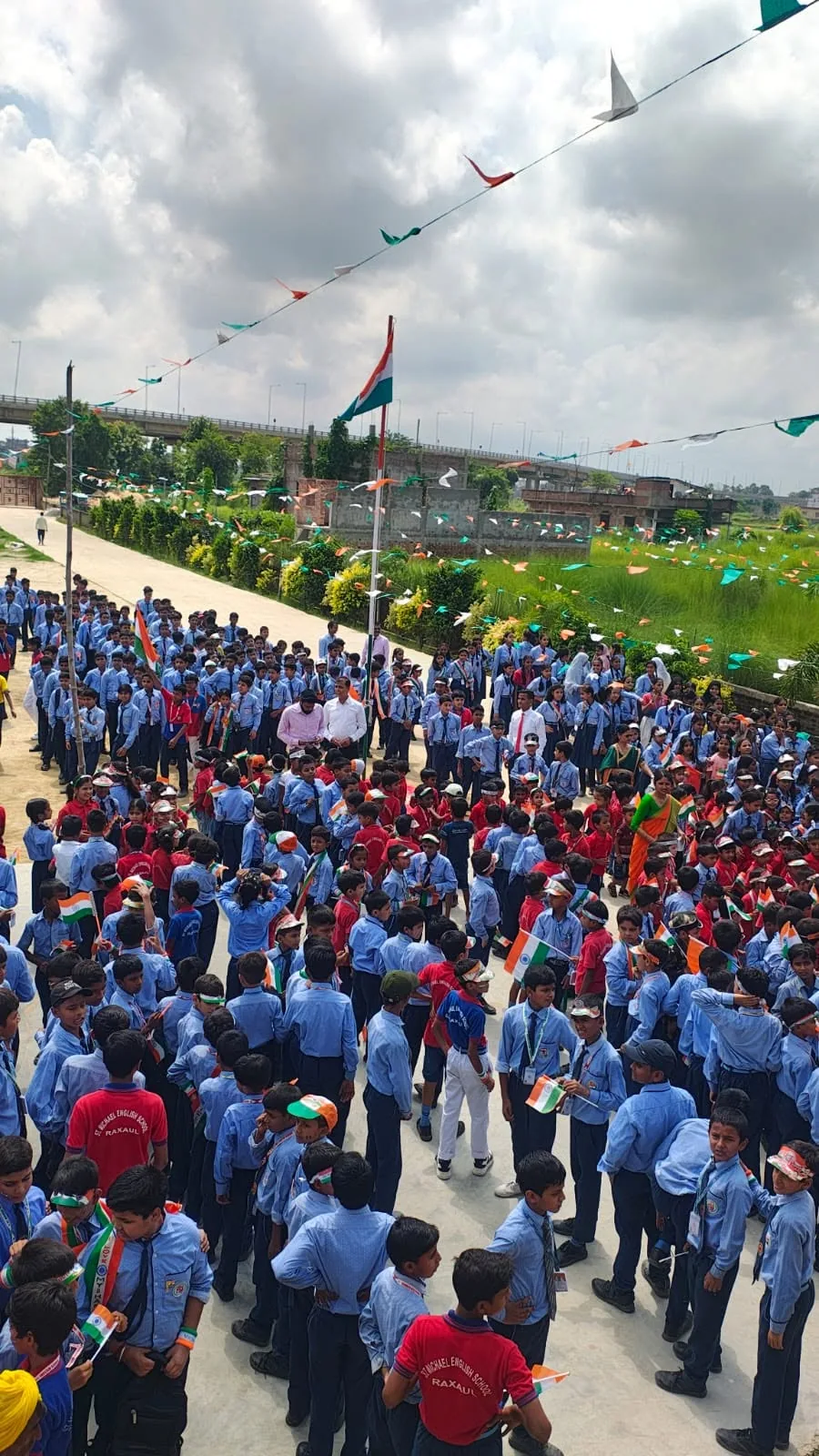 A wide view of the whole school assembled in the grounds beneath the national flag