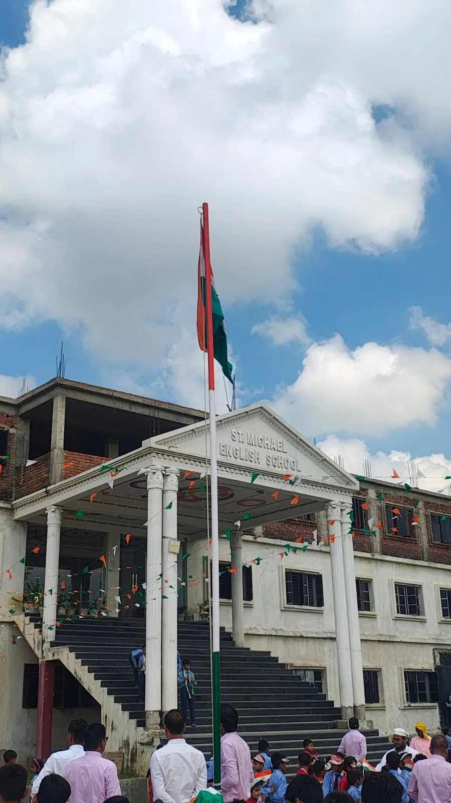 The main school building, a three-storey white building with a central staircase, with students assembled outside