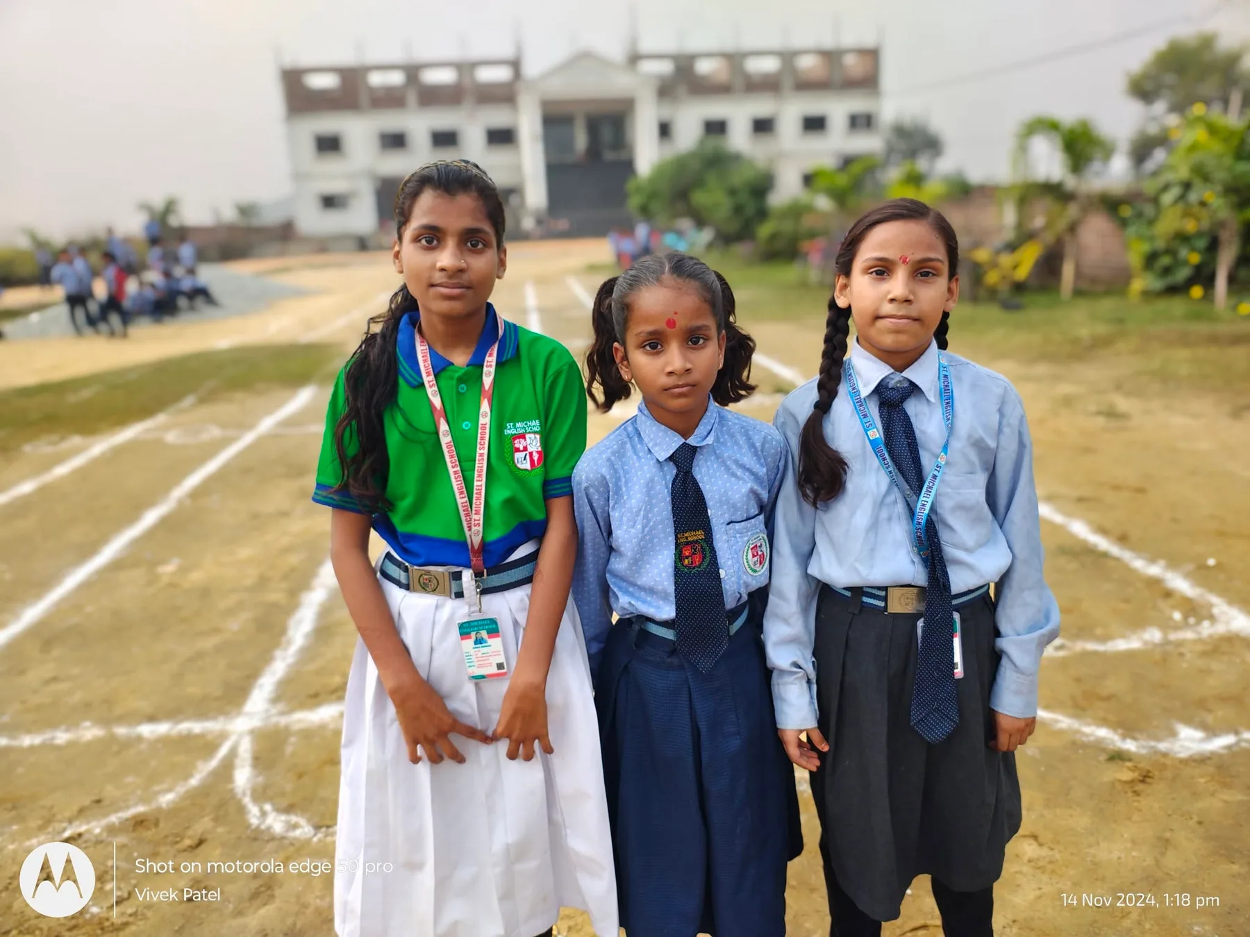 Three students in school uniform standing on the marked running track on sports day, with the school building behind them